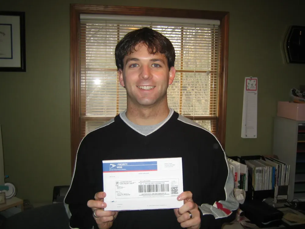 Ryan, one of the founders of MetaGeek, holding a USPS Priority Mail envelope indoors with office items visible in the background