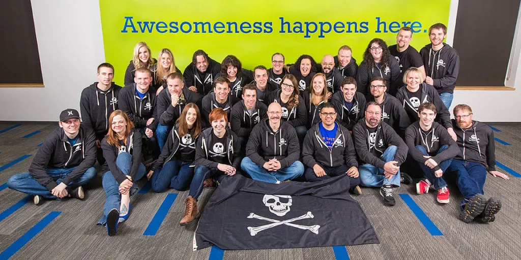 Group photo of the MetaGeek team posing indoors in front of a green and blue “Awesomeness happens here” banner, with many members wearing skull‑and‑crossbones hoodies and a pirate‑themed flag displayed at the front.